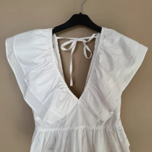 Vit klänning med volangdetaljer - Säljer en söt vit klänning med volangdetaljer och knytning i ryggen. Klänningen har en v-ringad design och är perfekt för sommarens varma dagar. Den är ärmlös och har en luftig passform.