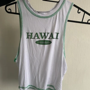 Vit linnetopp från Boohoo - Säljer en vit linnetopp från Boohoo med gröna detaljer och texten 'Hawaii Honolulu' på framsidan. Toppen har gröna sömmar runt ärmhål och halsringning. Perfekt för en avslappnad stil!