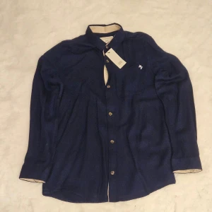 Mörkblå skjorta från River Island - Snygg mörkblå skjorta från River Island med klassisk krage och knappar framtill. Perfekt för en stilren look. Skjortan har långa ärmar och en liten broderad detalj på bröstet.