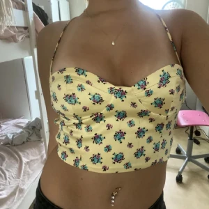 Gul blommig croppad satin linnetopp - Säljer en söt gul croppad linnetopp med blommigt mönster i blått och rosa. Toppen har smala axelband och en figurnära passform. Perfekt för sommardagar!