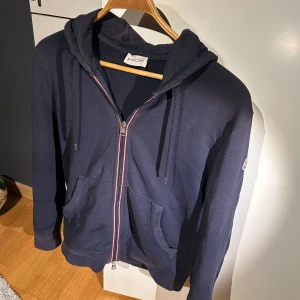 Mörkblå hoodie från Moncler - Säljer min fina mörkblå hoodie från Moncler med dragkedja och huva. Den är i använt men överlag bra skick. Enda synliga defekten är ett lagat hål på ena armbågen men inget som syns särskilt mycket. Äkta, och perfekt för en stilren samt bekväm look. Kan passa både S och M.