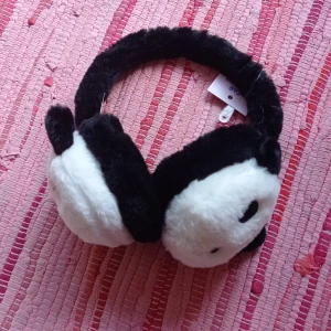 Svart och vit öronvärmare med pandamotiv - Mysiga öronvärmare i svart och vitt med gulligt pandamotiv. Perfekt för att hålla värmen under kalla dagar. Tillverkad i mjuk polyester för extra komfort. Aldrig använda. Vet ej vart de köptes