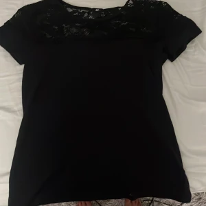 Svart topp med spetsdetaljer från H&M - Säljer en stilren svart topp från H&M med spetsdetaljer vid axlar och hals. Toppen har korta ärmar och en rund halsringning. Perfekt för en elegant look.