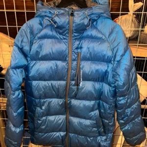 Blå dunjacka från McKinley - Säljer en blå dunjacka från McKinley med huva och dragkedja. Jackan är perfekt för kyliga dagar och har en stilren design med quiltade paneler. Den är långärmad och har justerbara snören vid huvan för extra skydd mot väder och vind.