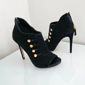 Svarta klackskor från Guess - Snygga svarta klackskor från Guess i mocka med öppen tå och dragkedja bak. Skorna har dekorativa guldknappar på sidan och en elegant klack. Perfekta för att ge en stilren touch till din outfit.