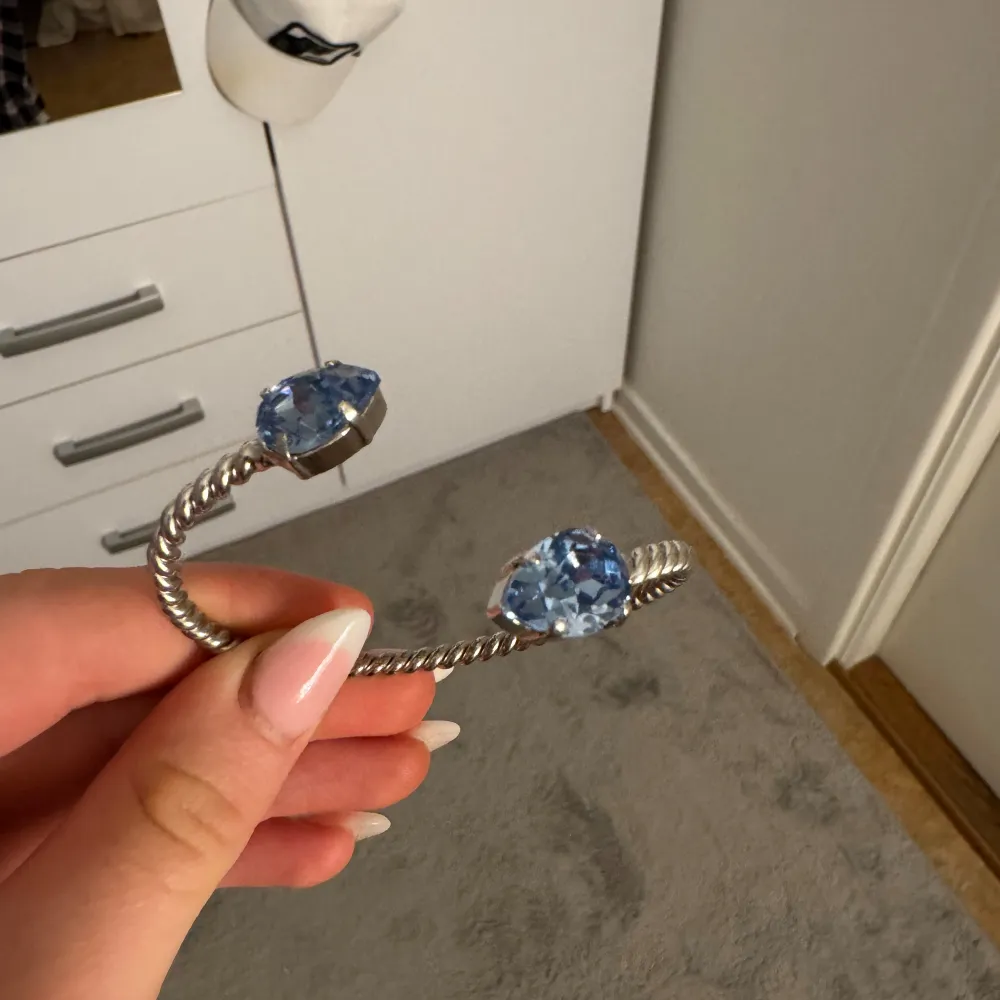 Snyggt silverfärgat armband med två stora blå stenar i ändarna. Armbandet har en vriden design som ger det en unik look. Perfekt för att lägga till en touch av färg och stil till din outfit.. Asusteet.