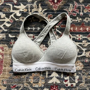 Grå sport bh från Calvin Klein - Säljer en snygg grå sport bh från Calvin Klein med klassisk logga på det elastiska bandet. Perfekt för en avslappnad stil. Toppen har en sportig design med korsade axelband. I bra skick, små defekter som syns på bilderna men annars i bra skick! Storlek xs 🩷