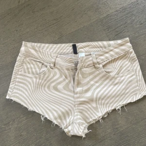 Beige  lågmidjade jeansshorts med vågigt mönster - Snygga beige shorts med ett unikt vågigt mönster. De har fransiga kanter och en dragkedja framtill. Perfekta för en avslappnad stil.