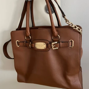 Brun axelväska från Michael Kors - Snygg brun axelväska från Michael Kors i skinn med gulddetaljer och en praktisk dragkedja. Väskan har dubbla handtag och en kedjedetalj på axelremmen. Perfekt för att bära dina dagliga nödvändigheter med stil.