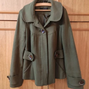 Grön vårkappa från Topshop - Supersöt grön kappa från Topshop med stora knappar och bred krage. Jackan har långa ärmar och dekorativa knappar vid ärmsluten. Perfekt för höst och vår! Superfint skick, skriv om du har några frågor!