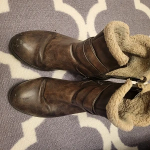 Bruna boots med foder - Säljer ett par bruna boots med mjukt foder och dekorativt bälte. De har en robust sula och dragkedja på sidan för enkel på- och avtagning. Perfekta för kyligare dagar.