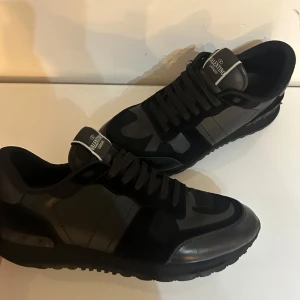Svarta sneakers från Valentino Garavani - Snygga svarta sneakers från Valentino Garavani med ett diskret camouflage-mönster. Skorna har snörning och en stilren design som passar perfekt för en trendig look.
