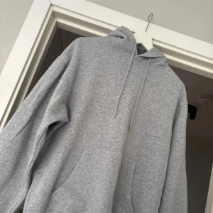 Grå hoodie - Säljer en klassisk grå hoodie med justerbar dragsko i huvan. Den har en bekväm passform och en stor ficka framtill. Perfekt för en avslappnad stil. Hör av dig vid intresse!
