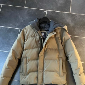 Olivgrön Canada Goose jacka - Säljer en olivgrön dunjacka från Canada Goose med huva och dragkedja. Jackan har en klassisk pufferdesign och en tydlig logotyp på ärmen. Perfekt för kalla dagar. Canada goose påsen ingår också. 