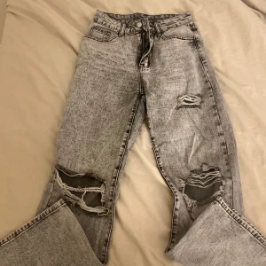 Grå jeans med slitningar - Säljer ett par grå jeans med coola slitningar på framsidan. De har en klassisk femficksdesign och en knappgylf. Perfekta för en avslappnad stil.