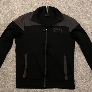 Svart och grå jacka från Boss - Snygg svart och grå tröja från Hugo Boss som är i perfekt skick, den är storlek M men passar lika bra från S till L. Tröjan är den gamla hugo boss modellen så den är inte så lätt att få tag på nuförtiden.