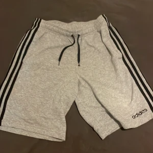 Grå shorts från Adidas - Snygga grå shorts från Adidas med klassiska svarta ränder längs sidorna. De har en bekväm resårmidja med snörning för justerbar passform. Perfekta för träning eller avslappnade dagar.