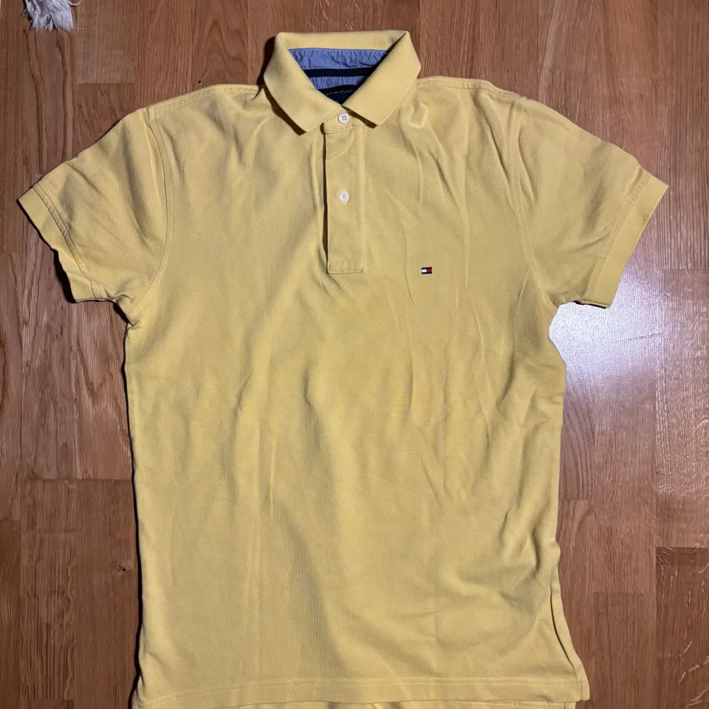 Säljer en snygg gul pikétröja från Tommy Hilfiger. Tröjan har en klassisk krage med två knappar och det ikoniska logotypmärket på bröstet. Perfekt för en stilren och avslappnad look.. Neuletakit & Villapaidat.