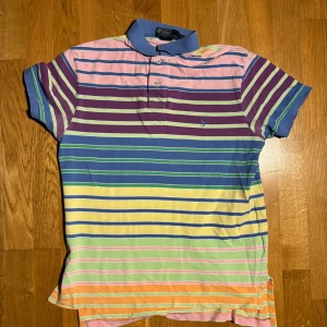 Flerfärgad randig pikétröja från Ralph Lauren - Säljer en färgglad pikétröja från Ralph Lauren med ränder i rosa, lila, blå, gul och grön. Tröjan har korta ärmar och en klassisk krage med knappar. Perfekt för en avslappnad stil med en touch av färg!