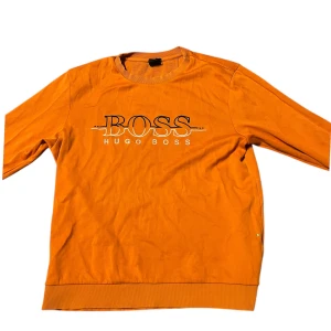 Orange tröja från Hugo Boss - Snygg orange tröja från Hugo Boss med stort tryck på framsidan. Tröjan har en rund halsringning och är perfekt för en avslappnad stil. Passar bra till jeans eller chinos.