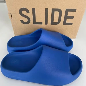Yeezy Slide azure från Adidas - Säljer ett par blå YZY Slide från Adidas. Dessa sandaler har en modern design med en bred rem över foten och en räfflad sula för extra grepp. Perfekta för en avslappnad stil. Perfect inför sommaren har dock en skrämma vid vänster fot.