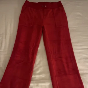 Röda byxor från Juicy Couture - Snygga röda byxor från Juicy Couture med fickor och knappar bak. Byxorna har en loose passform och är tillverkade i ett mjukt material som ger en bekväm känsla. Perfekta för en avslappnad stil. Tyvärr så har snörena åkt ur och gick inte att få tillbaka men annars är byxorna helt fria från defekter och är endast använda ett fåtal gånger❤️