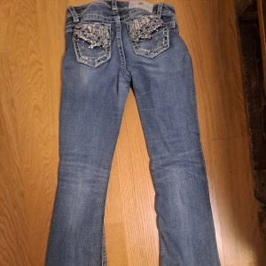 Blå jeans med paljettdetaljer - Snygga blå jeans med bootcut-stil och glittriga paljettdetaljer på bakfickorna. Perfekta för att ge en extra touch till din outfit. De har en lågmidjad passform och är tillverkade i klassiskt jeanstyg.