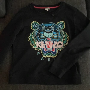 Kenzo tröja  - Säljer en svart tröja från Kenzo med ett coolt tigertryck i blått och rosa på framsidan. Tröjan har långa ärmar och en rund halsringning. Perfekt för dig som vill ha en stilren men ändå iögonfallande look. 🐯