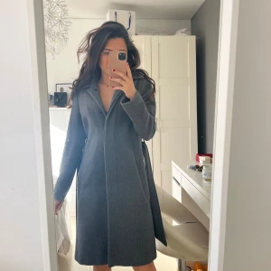 Grå kappa från Filippa K - Snygg grå ullkappa från Filippa K i storlek XS. Den har en klassisk design med knappar framtill och ett bälte i midjan för en elegant siluett. Perfekt för kyligare dagar med sin långa ärm och stilrena look.