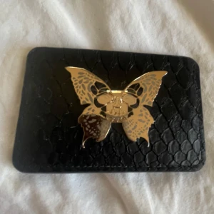 Svart plånbok med fjärilsdetalj - Snygg svart plånbok från Lecacci med en unik fjärilsdetalj i guld på framsidan. Plånboken har ett elegant mönster som påminner om ormskinn och flera fack för kort. Perfekt för att hålla ordning på dina kort med stil.