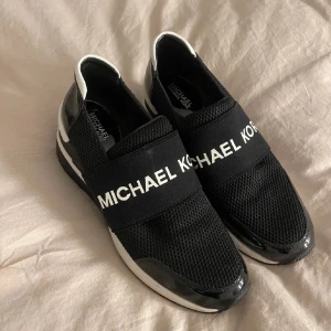 Svarta sneakers från Michael Kors - Snygga svarta sneakers från Michael Kors med vit logga på ett brett band över foten. Skorna har en stilren design med svarta och vita detaljer samt en bekväm passform. Perfekta för en trendig look.