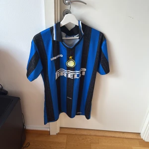 Blå och svart randig fotbollströja från Umbro - Säljer en klassisk blå och svart randig fotbollströja från Umbro med korta ärmar. Tröjan har ett tryck med 'Ronaldo' och nummer 10 på ryggen. Perfekt för fotbollsfans och samlare!