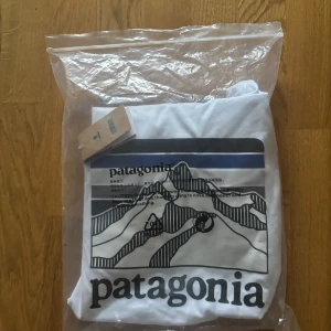 Vit t-shirt från Patagonia - Säljer en vit t-shirt från Patagonia med ett stilrent tryck av berg och logotyp på baksidan. T-shirten har en normal passform och är tillverkad av återvunnen bomull. Perfekt för en avslappnad stil.