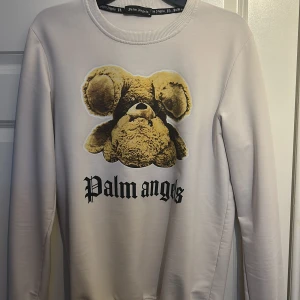 Vit tröja från Palm Angels med nallemotiv - Säljer en vit tröja från Palm Angels med ett unikt nallemotiv på framsidan och text på både fram- och baksidan. Tröjan har långa ärmar och en rund halsringning. Perfekt för dig som vill ha något stilrent men ändå iögonfallande.