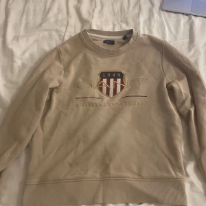 Beige sweatshirt från GANT - Säljer en beige sweatshirt från GANT med ett broderat emblem och texten '1949 American Sportswear' på framsidan. Tröjan har långa ärmar och ribbade muddar vid ärmslut och nederkant. Perfekt för en avslappnad stil.