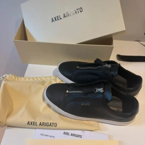 Svarta sneakers från Axel Arigato med dragkedja - Snygga svarta sneakers från Axel Arigato i skinn med vit sula och dragkedja framtill istället för klassisk snörning. Minimalistisk design med diskret logga på sidan. Perfekta för dig som gillar stilrena och moderna skor. Använd några gånger men fortfarande jätte fint skick, allt på bilderna medföljer! Vid frågor är de bara att skriva!😀