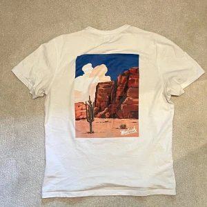 Vit t-shirt med tryck från Woolrich - Vit t-shirt från Woolrich med liten logga på bröstet och stort färgglatt tryck av ökenlandskap med kaktus och röda klippor på ryggen. Den perfekta sommar tirshan! Vid minsta fråga eller fundering hör av er🤝 OBS! Slutet tryck på ryggen