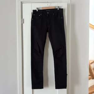 Svarta jeans från Nudie Jeans - Säljer ett par svarta jeans från Nudie Jeans med klassisk femficksdesign och raka ben. Jeansen har knappgylf och är tillverkade i ett mjukt bomullstyg med lite stretch. Perfekta för dig som gillar stilrena och tidlösa byxor.