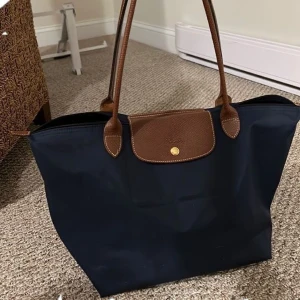 Marinblå handväska från Longchamp - Säljer en klassisk marinblå handväska från Longchamp med bruna skinnhandtag och lock. Väskan har guldfärgade metalldetaljer och dragkedja. Perfekt storlek för vardagsbruk och rymmer det mesta du behöver.