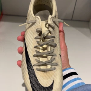 Beige Nike mercurial fotbollsskor med svart swoosh - Säljer ett par beigea Nike fotbollsskor med svart swoosh på sidan. Skorna har snörning och är designade för att ge bra grepp på planen med sin mönstrade sula. Perfekta för dig som vill ha stilrena och funktionella skor till fotbollsträningen.