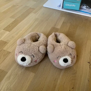 Bruna tofflor med björnansikte - Supergulliga bruna tofflor med mjukt plyschmaterial och söta björnansikten framtill. Perfekta för att hålla fötterna varma hemma, med små öron och broderade detaljer som ger en lekfull look.
