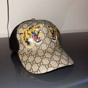 Gucci keps med tigerbrodyr - Snygg keps från Gucci med klassiskt GG-mönster i beige och brunt. Framsidan har broderade tigrar och baksidan är i svart mesh med justerbart spänne och röd-grön detalj. Perfekt accessoar för en trendig look.