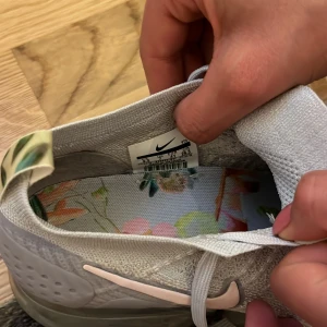 Nike Air Vapormax Flyknit sneakers i ljusgrått - Säljer ett par Nike Air Vapormax Flyknit sneakers i ljusgrå färg med stickad ovandel och genomskinlig sula. Skorna har vita snören, rosa swoosh och färgglada detaljer på hälen samt blommigt mönstrad innersula.