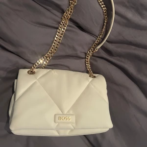 Vit axelväska från BOSS - Säljer en stilren vit axelväska från BOSS med guldfärgad kedja. Väskan har en elegant look med BOSS-logga framtill och passar perfekt när du vill lyfta din outfit.