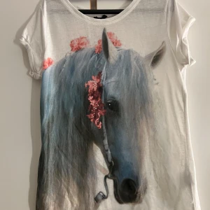 Vit t-shirt med hästmotiv  - Vit t-shirt från Kappahl med stort tryck av en vit häst med rosa blommor i manen. T-shirten har rund halsringning och korta ärmar. Tillverkad i 100% polyester, perfekt för dig som gillar djurmotiv och unika prints.