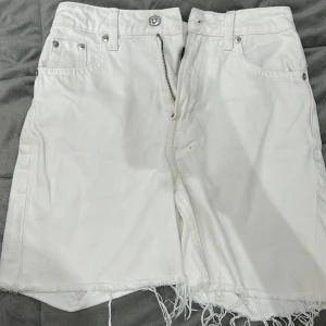 Vit jeansshort från Divided - Säljer en vit jeansshort från Divided med råa kanter och klassiska fickor fram. Kjolen har dragkedja och knapp framtill samt bälteshällor. Perfekt för en trendig och avslappnad look.