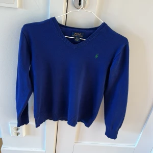 Blå v-ringad tröja från Polo Ralph Lauren - Säljer en klassisk blå v-ringad tröja från Polo Ralph Lauren med den ikoniska gröna broderade loggan på bröstet. Tröjan har långa ärmar och är tillverkad i mjukt material som passar perfekt till många tillfällen.