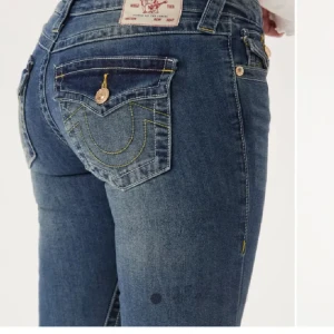 Blå bootcut jeans från True Religion - Säljer ett par klassiska blå jeans från True Religion med bootcut-passform. Jeansen har dekorativa sömmar och guldiga knappar på bakfickorna samt en tydlig logotyp-patch baktill. Perfekta för dig som gillar en tidlös jeanslook.