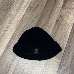Svart stickad mössa från Parajumpers - Svart stickad mössa från Parajumpers med ribbad uppvikning och diskret logotyp framtill. Klassisk modell med flätstickat mönster som passar perfekt till kyliga dagar. Pris kan diskuteras vid snabb affär. 