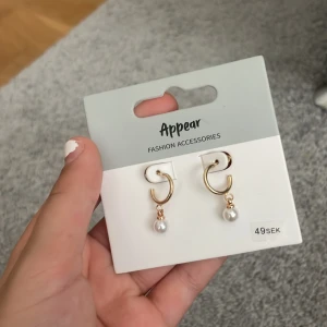 Guldiga örhängen med pärldetalj från Appear - Snygga guldiga örhängen med en hängande vit pärla. En guldig cirkelform och fin pärla. Perfekta för att lyfta din stil med en trendig touch.